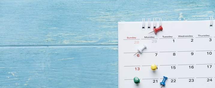 An einer blauen Holzwand hängt ein Monatskalender mit einigen Pinsteckern.