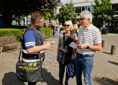 Ein Mann mit einer Tasche, auf der Impflotsen steht, spricht mit einem älteren Paar.