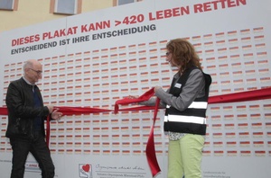 Ein Mann und eine Frau öffnen eine Schleife vor einem großen Plakat.